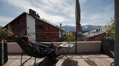 Young beautiful woman basking in the sun lying in a chair in a plaid on terrace with mountain view