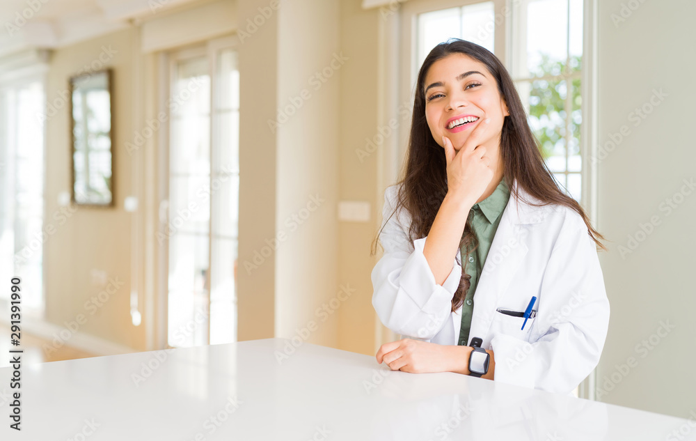 Young woman wearing medical coat at the clinic as therapist or doctor looking confident at the camera with smile with crossed arms and hand raised on chin. Thinking positive. Young woman wearing medical coat at the clinic as therapist or doctor looking confident at the camera with smile with crossed arms and hand raised on chin. Thinking positive.