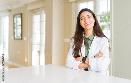 Young woman wearing medical coat at the clinic as therapist or doctor happy face smiling with crossed arms looking at the camera. Positive person.
