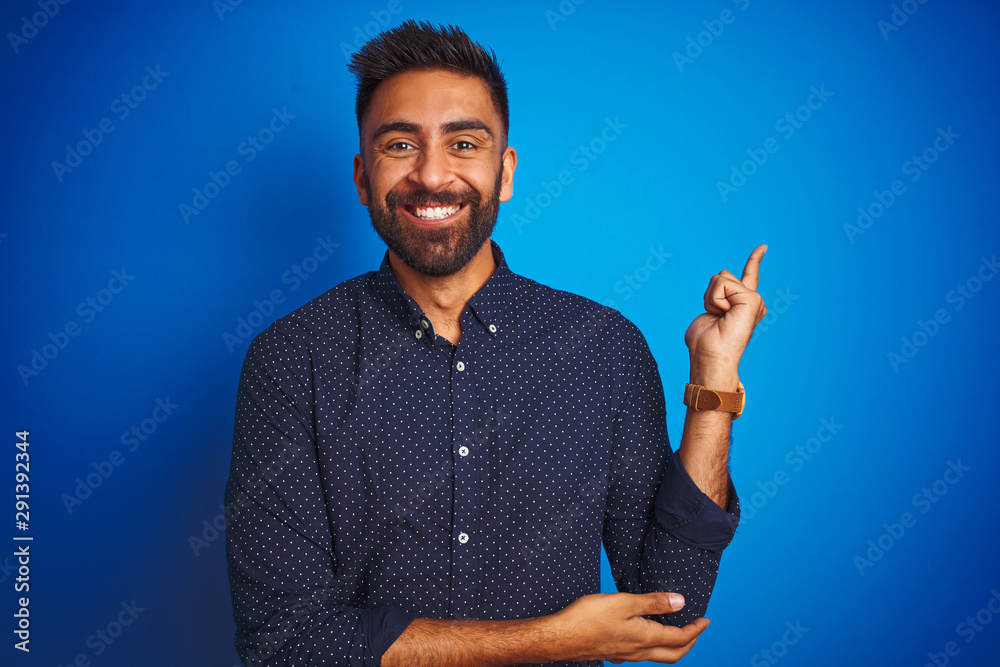 Young indian elegant man wearing shirt standing over isolated blue background with a big smile on face, pointing with hand and finger to the side looking at the camera. - obrazy, fototapety, plakaty 