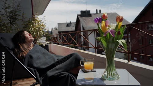 Happy smiling young woman in plaid enjoying spring sun on terrace. Bouquet of colorful tulips and cup of orange juice stand next on table