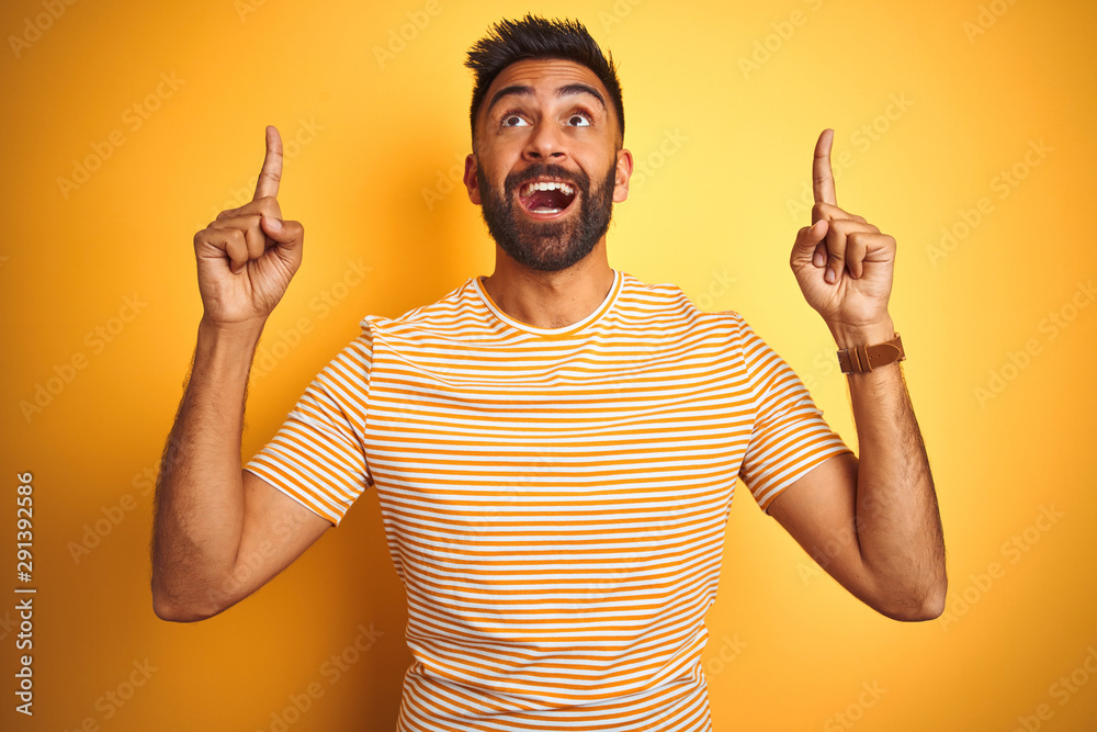 Young indian man wearing t-shirt standing over isolated yellow background smiling amazed and surprised and pointing up with fingers and raised arms.