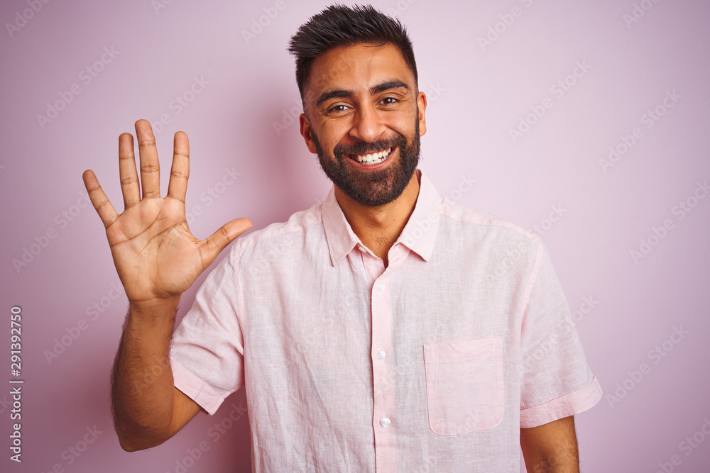 Young indian man wearing casual shirt standing over isolated pink background showing and pointing up with fingers number five while smiling confident and happy.