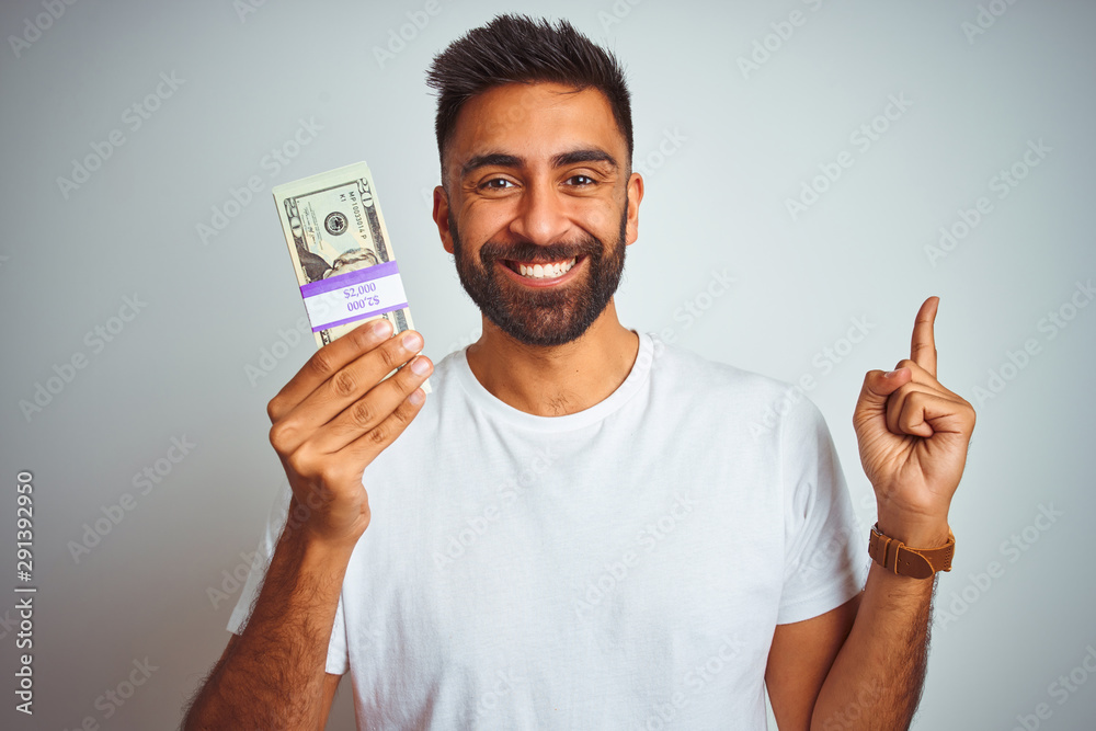 Young indian man holding dollars standing over isolated white background very happy pointing with hand and finger to the side Young indian man holding dollars standing over isolated white background very happy pointing with hand and finger to the side