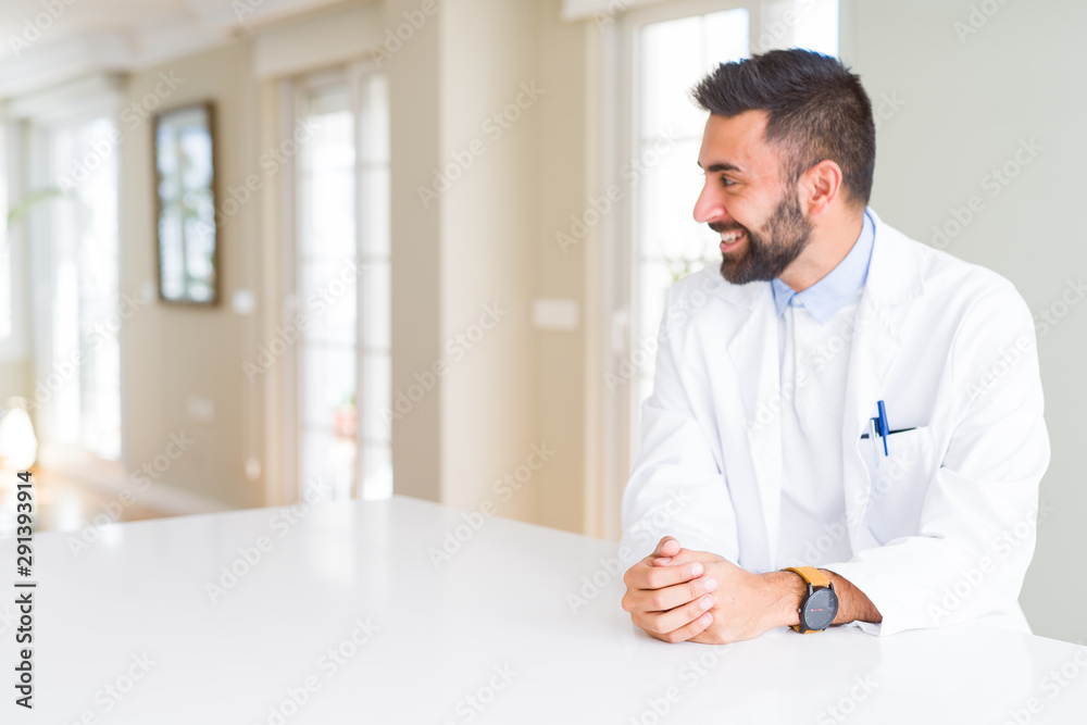 Handsome hispanic doctor or therapist man wearing medical coat at the clinic looking away to side with smile on face, natural expression. Laughing confident.