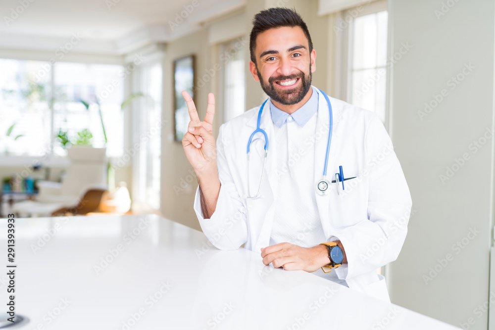 Handsome hispanic doctor man wearing stethoscope at the clinic smiling with happy face winking at the camera doing victory sign. Number two. Handsome hispanic doctor man wearing stethoscope at the clinic smiling with happy face winking at the camera doing victory sign. Number two.