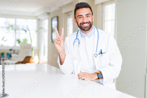 Handsome hispanic doctor man wearing stethoscope at the clinic smiling with happy face winking at the camera doing victory sign. Number two.
