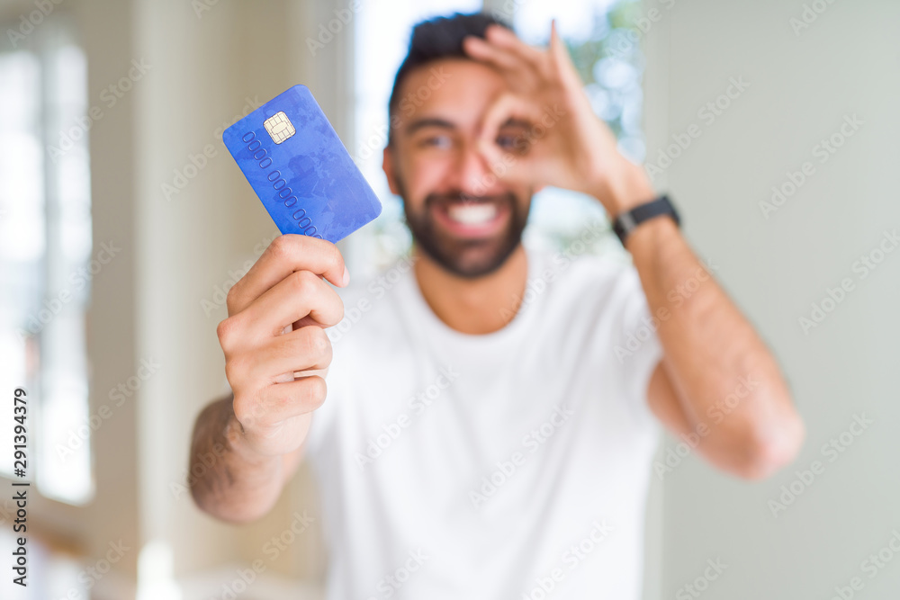 Handsome hispanic man holding credit card with happy face smiling doing ok sign with hand on eye looking through fingers - obrazy, fototapety, plakaty 