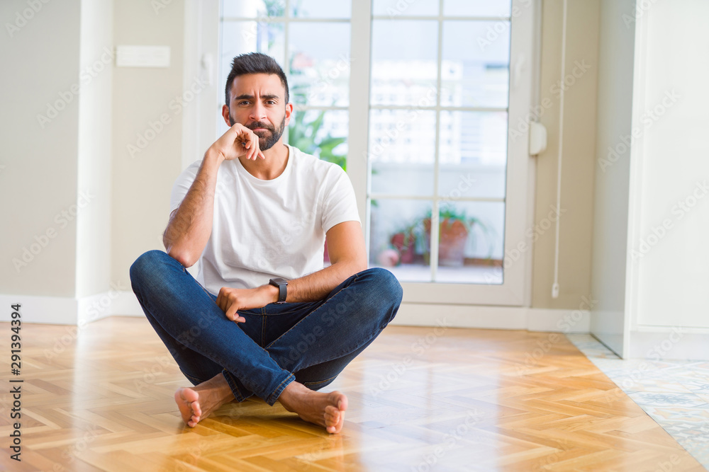 Handsome hispanic man wearing casual t-shirt sitting on the floor at home with hand on chin thinking about question, pensive expression. Smiling with thoughtful face. Doubt concept.