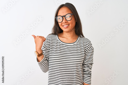 Young chinese woman wearing striped t-shirt and glasses over isolated white background smiling with happy face looking and pointing to the side with thumb up.