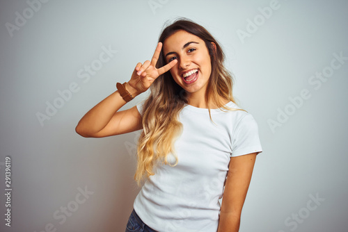 Young beautiful woman wearing casual white t-shirt over isolated background Doing peace symbol with fingers over face, smiling cheerful showing victory