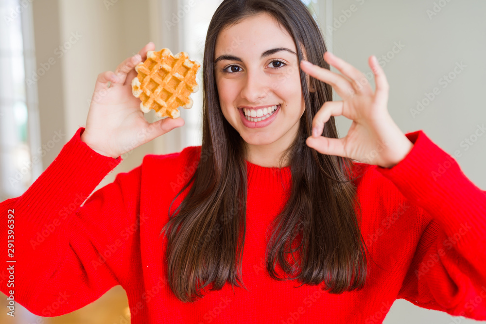 Beautiful young woman eating sweet belgian waffle pastry doing ok sign with fingers, excellent symbol