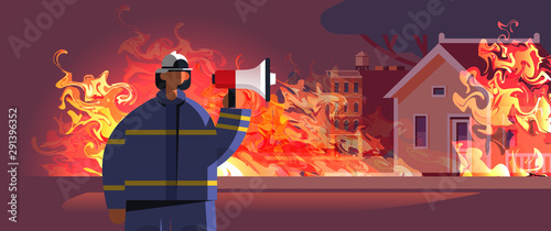brave fireman holding loudspeaker firefighter in uniform and helmet firefighting emergency service extinguishing fire concept burning house exterior orange flame background portrait