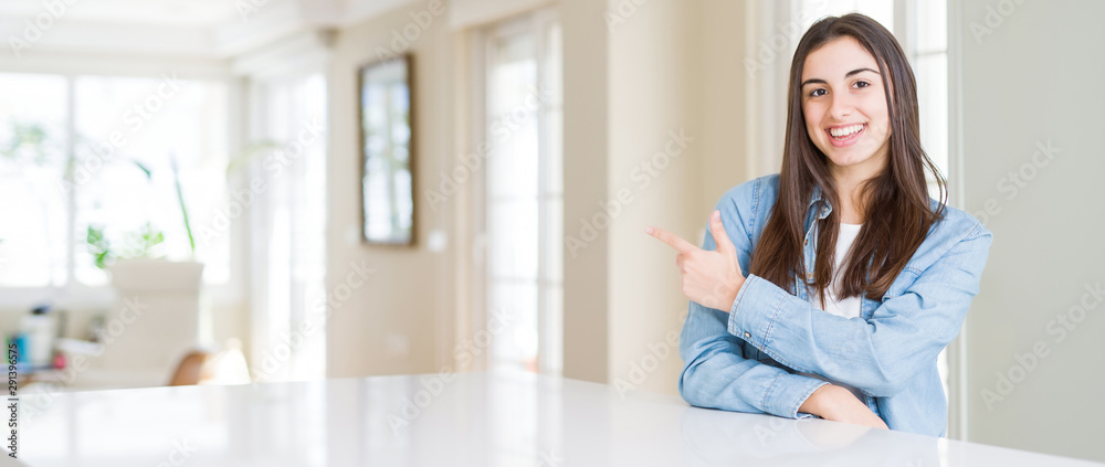 Wide angle picture of beautiful young woman sitting on white table at home cheerful with a smile of face pointing with hand and finger up to the side with happy and natural expression on face Wide angle picture of beautiful young woman sitting on white table at home cheerful with a smile of face pointing with hand and finger up to the side with happy and natural expression on face