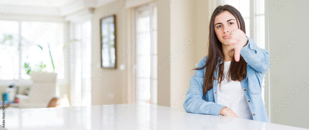 Wide angle picture of beautiful young woman sitting on white table at home looking unhappy and angry showing rejection and negative with thumbs down gesture. Bad expression.