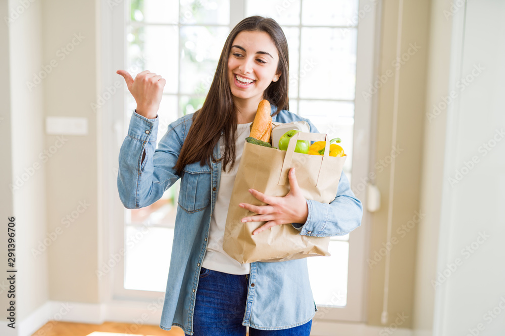 Beautiful young woman holding paper bag full of healthy groceries pointing and showing with thumb up to the side with happy face smiling Beautiful young woman holding paper bag full of healthy groceries pointing and showing with thumb up to the side with happy face smiling