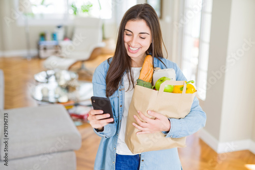 Young woman holding a paper bag full of fresh groceries and using smartphone app for supermarket delivery photo