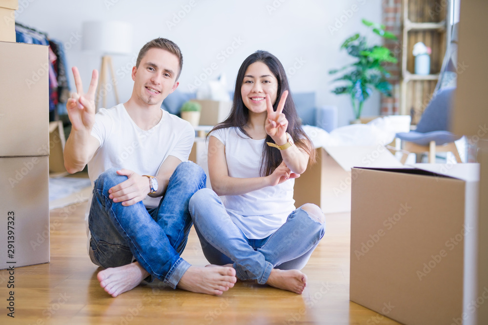 Young beautiful couple sitting on the floor at new home around cardboard boxes smiling with happy face winking at the camera doing victory sign. Number two. Young beautiful couple sitting on the floor at new home around cardboard boxes smiling with happy face winking at the camera doing victory sign. Number two.