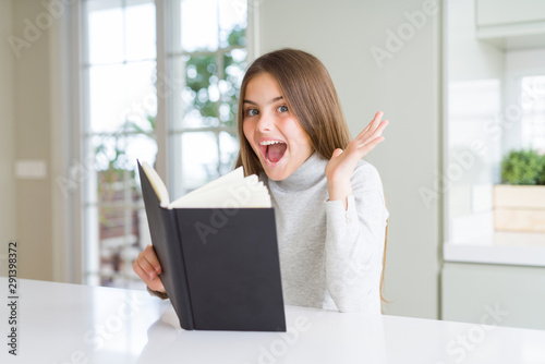 Beautiful young girl reading a book at home very happy and excited, winner expression celebrating victory screaming with big smile and raised hands