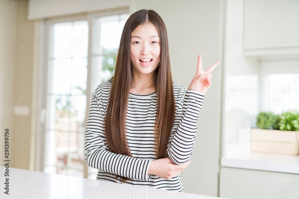 Beautiful Asian woman wearing stripes sweater smiling with happy face winking at the camera doing victory sign. Number two.