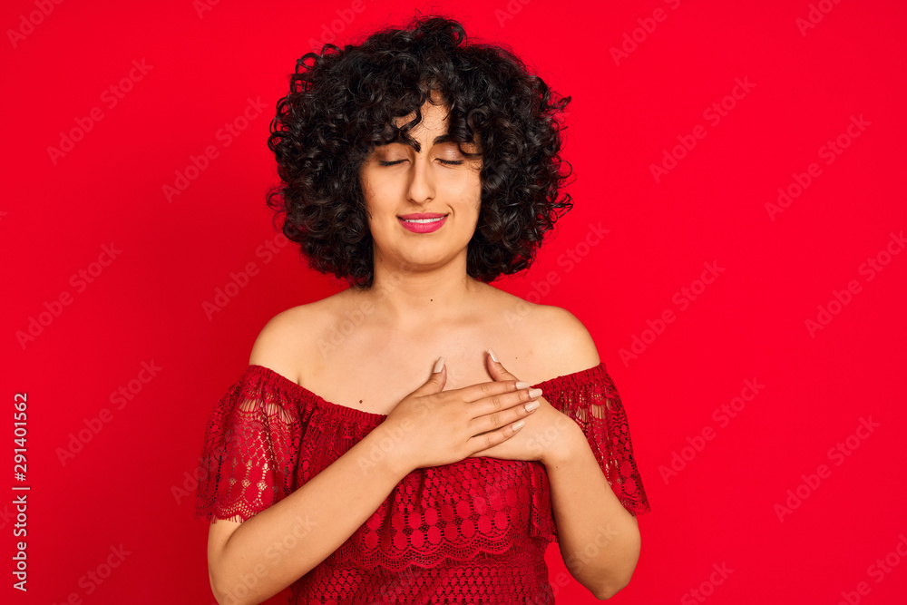 Young arab woman with curly hair wearing casual dress over isolated red background smiling with hands on chest with closed eyes and grateful gesture on face. Health concept. Young arab woman with curly hair wearing casual dress over isolated red background smiling with hands on chest with closed eyes and grateful gesture on face. Health concept.