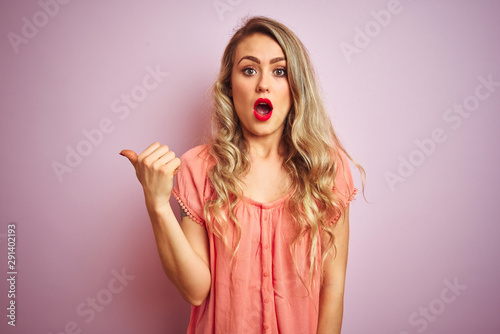 Young beautiful woman wearing t-shirt standing over pink isolated background Surprised pointing with hand finger to the side, open mouth amazed expression.