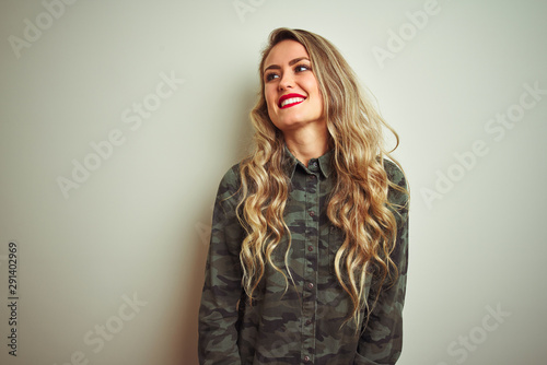 Young beautiful woman wearing military camouflage shirt over white isolated background looking away to side with smile on face, natural expression. Laughing confident.