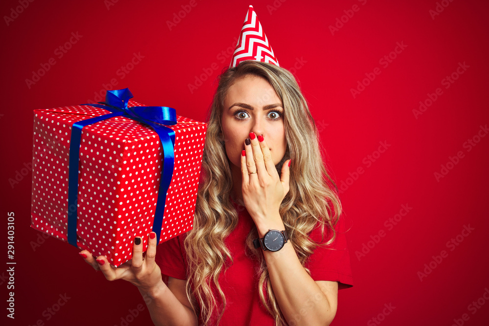 Young beautiful woman holding bitrhday gift over red isolated background cover mouth with hand shocked with shame for mistake, expression of fear, scared in silence, secret concept