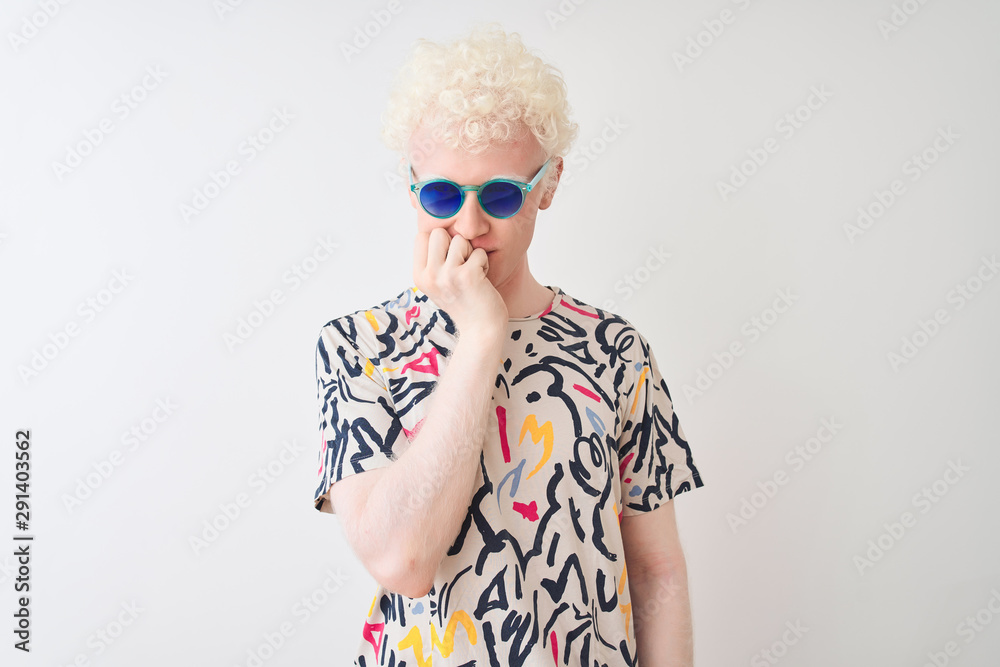 Young albino blond man wearing colorful t-shirt and sunglasses over isolated red background looking stressed and nervous with hands on mouth biting nails. Anxiety problem. Young albino blond man wearing colorful t-shirt and sunglasses over isolated red background looking stressed and nervous with hands on mouth biting nails. Anxiety problem.