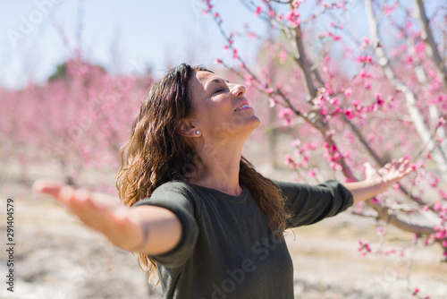 Beautiful middle age woman in the middle of pink peach flowers and trees smiling cheerful with open arms enjoying sunbathe on sunny day