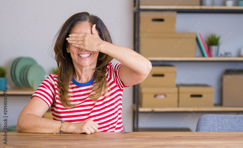 Middle age senior woman sitting at the table at home smiling and laughing with hand on face covering eyes for surprise. Blind concept. Middle age senior woman sitting at the table at home smiling and laughing with hand on face covering eyes for surprise. Blind concept.
