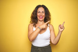 Middle age senior woman with curly hair standing over yellow isolated background smiling and looking at the camera pointing with two hands and fingers to the side.