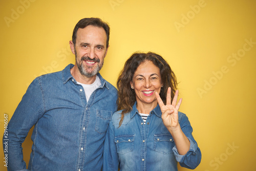 Beautiful middle age couple together standing over isolated yellow background showing and pointing up with fingers number four while smiling confident and happy.