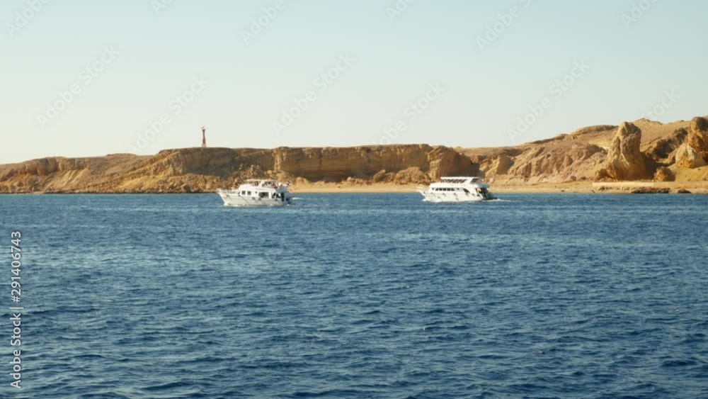 Small boats and yachts sailing along waves in blue sea near coastline with desert and stones at background Small boats and yachts sailing along waves in blue sea near coastline with desert and stones at background