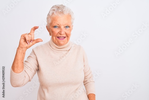 Senior grey-haired woman wearing turtleneck sweater standing over isolated white background smiling and confident gesturing with hand doing small size sign with fingers looking and the camera