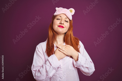 Young beautiful redhead woman wearing pajama and mask over purple isolated background smiling with hands on chest with closed eyes and grateful gesture on face. Health concept.