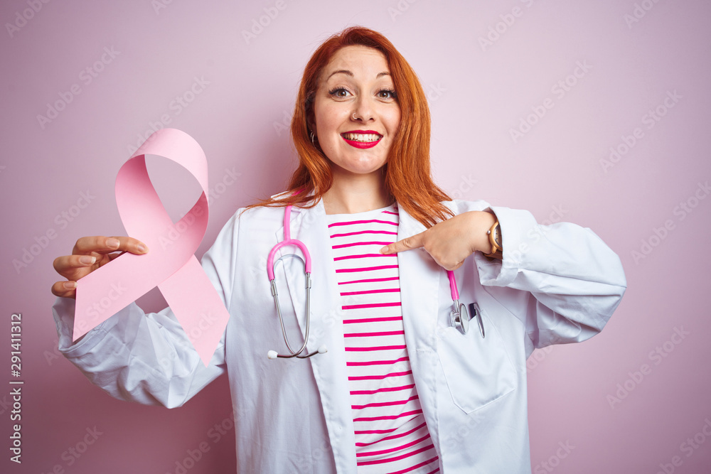 Young redhead doctor woman holding cancer ribbon over pink isolated background with surprise face pointing finger to himself Young redhead doctor woman holding cancer ribbon over pink isolated background with surprise face pointing finger to himself