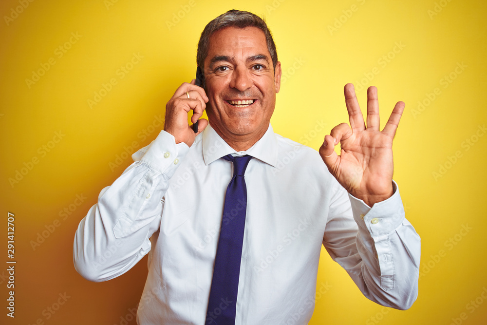 Middle age businessman talking on the smartphone over isolated yellow background doing ok sign with fingers, excellent symbol Middle age businessman talking on the smartphone over isolated yellow background doing ok sign with fingers, excellent symbol