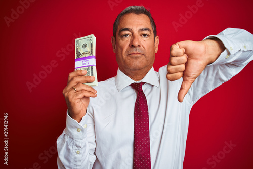 Handsome middle age businessman holding dollars over isolated red background with angry face, negative sign showing dislike with thumbs down, rejection concept