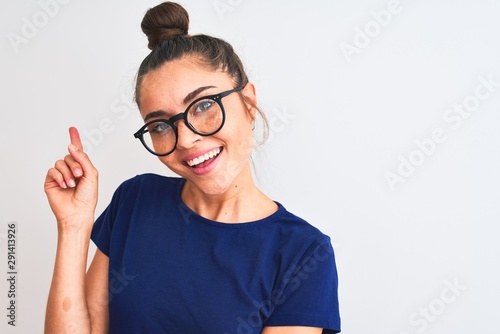 Beautiful woman with bun wearing blue t-shirt and glasses over isolated white background very happy pointing with hand and finger to the side