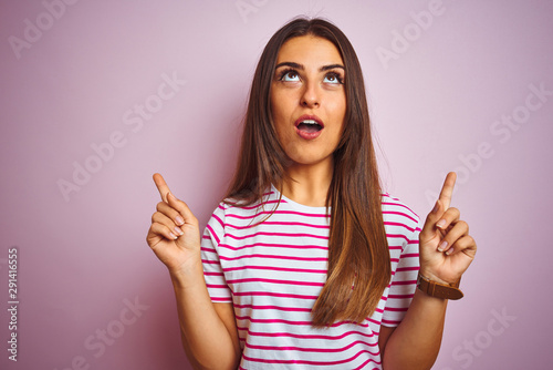 Young beautiful woman wearing striped t-shirt standing over isolated pink background amazed and surprised looking up and pointing with fingers and raised arms.