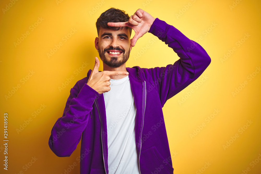 Young man with tattoo wearing sport purple sweatshirt over isolated yellow background smiling making frame with hands and fingers with happy face. Creativity and photography concept. Young man with tattoo wearing sport purple sweatshirt over isolated yellow background smiling making frame with hands and fingers with happy face. Creativity and photography concept.