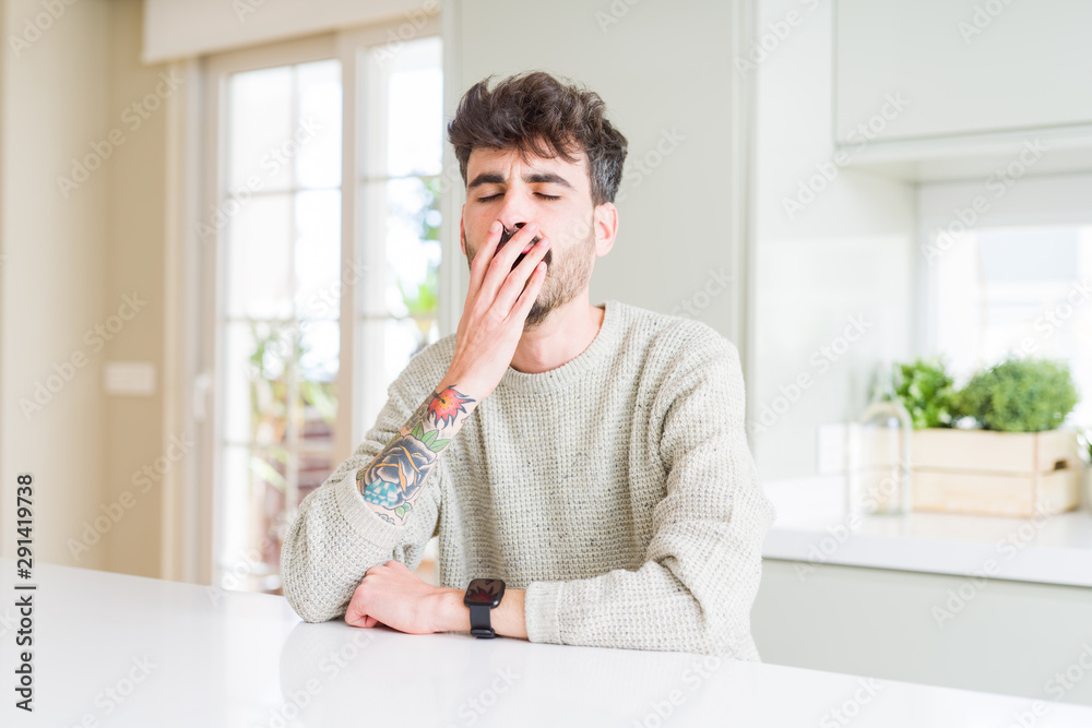 Young man wearing casual sweater sitting on white table bored yawning tired covering mouth with hand. Restless and sleepiness. Young man wearing casual sweater sitting on white table bored yawning tired covering mouth with hand. Restless and sleepiness.