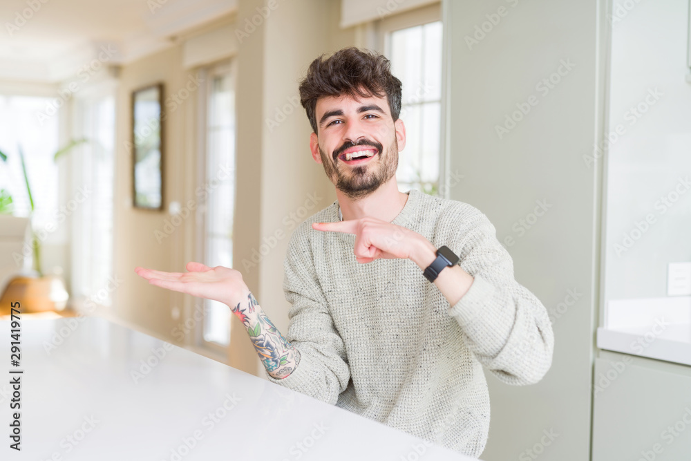 Young man wearing casual sweater sitting on white table amazed and smiling to the camera while presenting with hand and pointing with finger. Young man wearing casual sweater sitting on white table amazed and smiling to the camera while presenting with hand and pointing with finger.