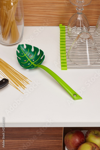 Subject shot of green spaghetti spoon in the form of a manstera leaf surrounded with spaghetti in a jar, a spaghetti wisp on the table, a dish rack with clean glasses on it and apples in the white bow photo