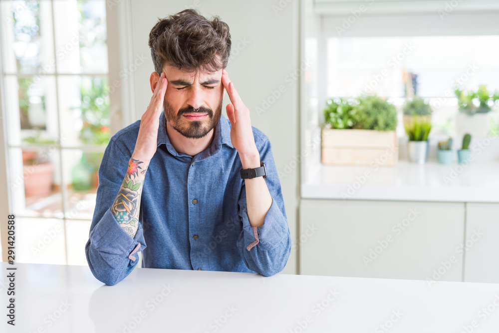 Young man wearing casual shirt sitting on white table with hand on head for pain in head because stress. Suffering migraine.