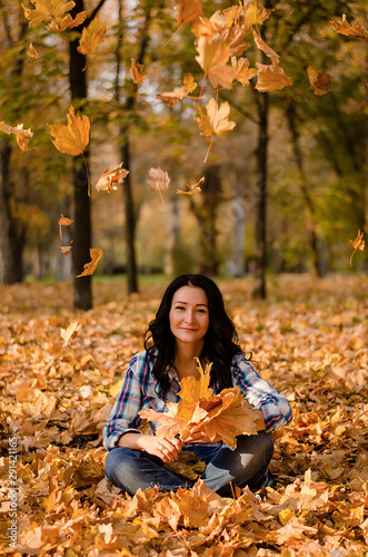 Beautiful woman sitting on the ground on yellow leaves in park. Leisure time on warm autumn day