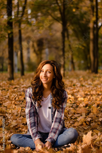 Beautiful girl sitting on the ground on yellow leaves in park. Leisure time on warm autumn day