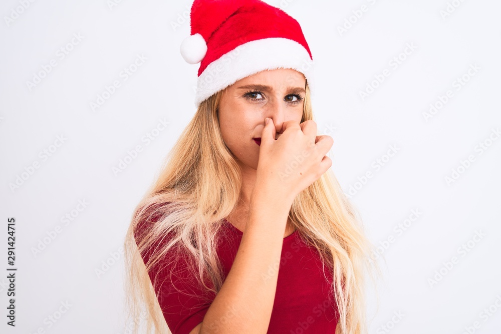 Young beautiful woman wearing Christmas Santa hat over isolated white background smelling something stinky and disgusting, intolerable smell, holding breath with fingers on nose. Bad smells concept. Young beautiful woman wearing Christmas Santa hat over isolated white background smelling something stinky and disgusting, intolerable smell, holding breath with fingers on nose. Bad smells concept.