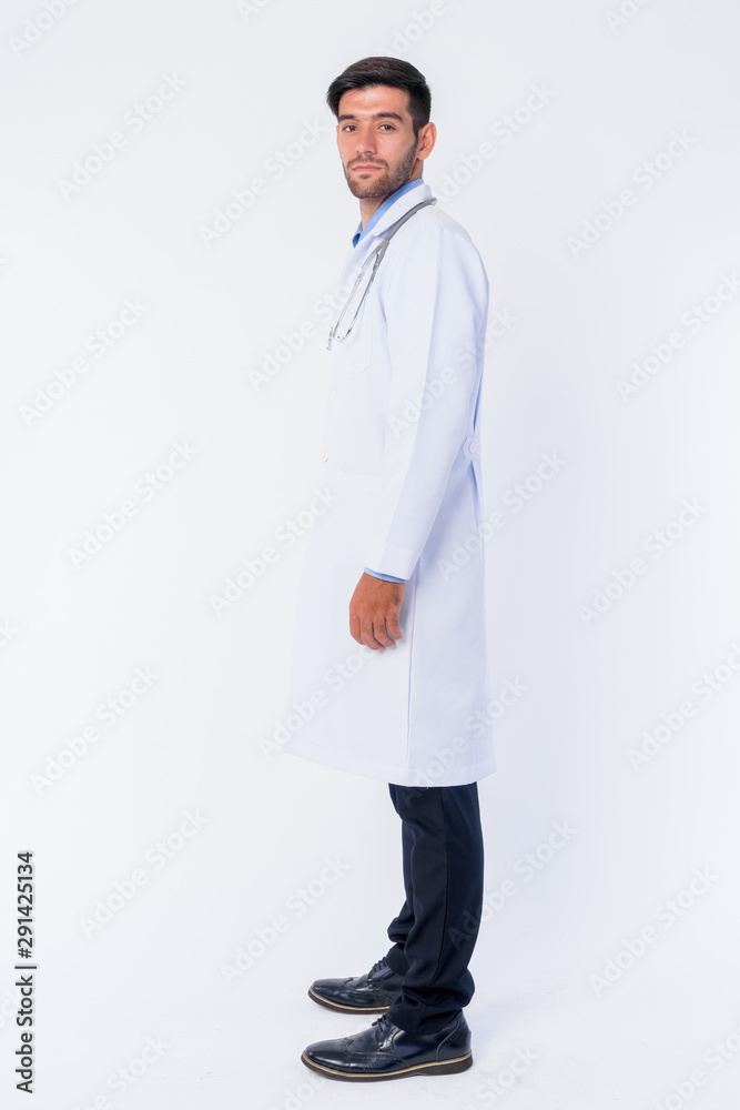 Full body shot profile view of young bearded Persian man doctor looking at camera